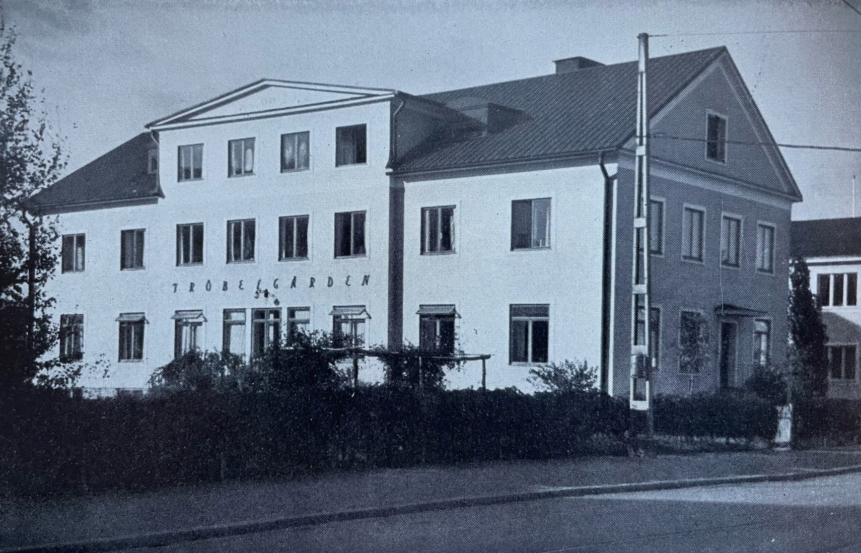 Äldre svartvitt foto visar en stod byggnad vid en gata. På byggnadens ena väg står texten "Fröbelgården".
